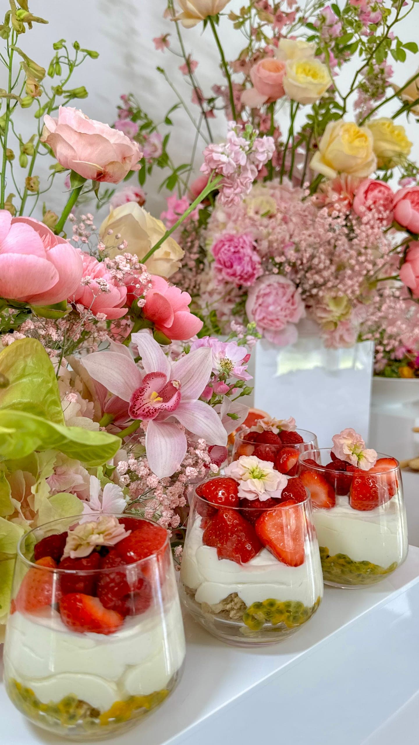 Dessertgläser mit Passionsfrucht, Knuspermüsli, Creme und Erdbeeren, dahinter pastellfarbene Blumengestecke