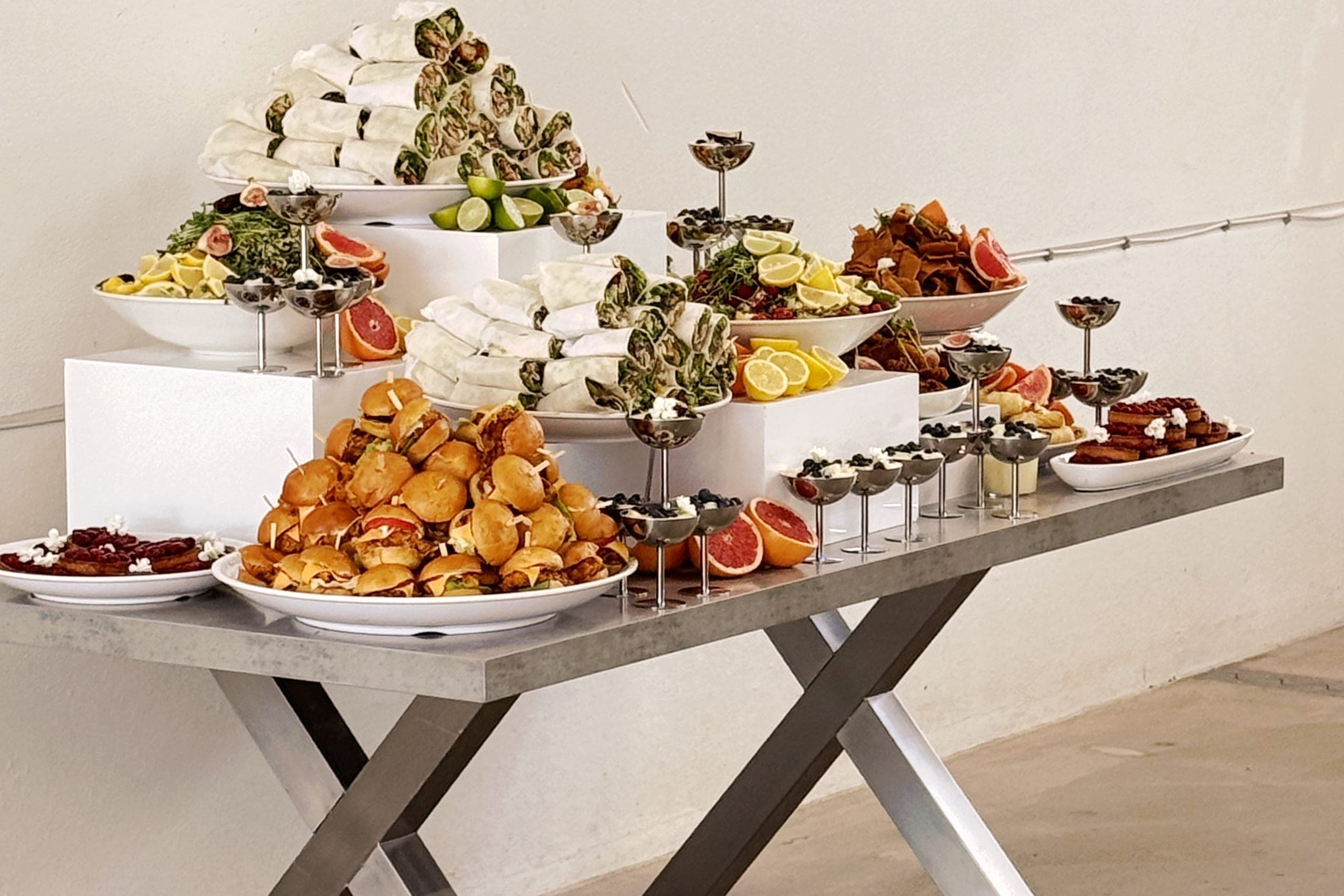 Firmenevent-Buffet auf weißen Sockeln: Wrap-Pyramiden, Mini-Burger, Kräutersalat mit Grapefruit und Zitrone, Dessertgläser mit Blaubeeren auf einem grauen Tisch