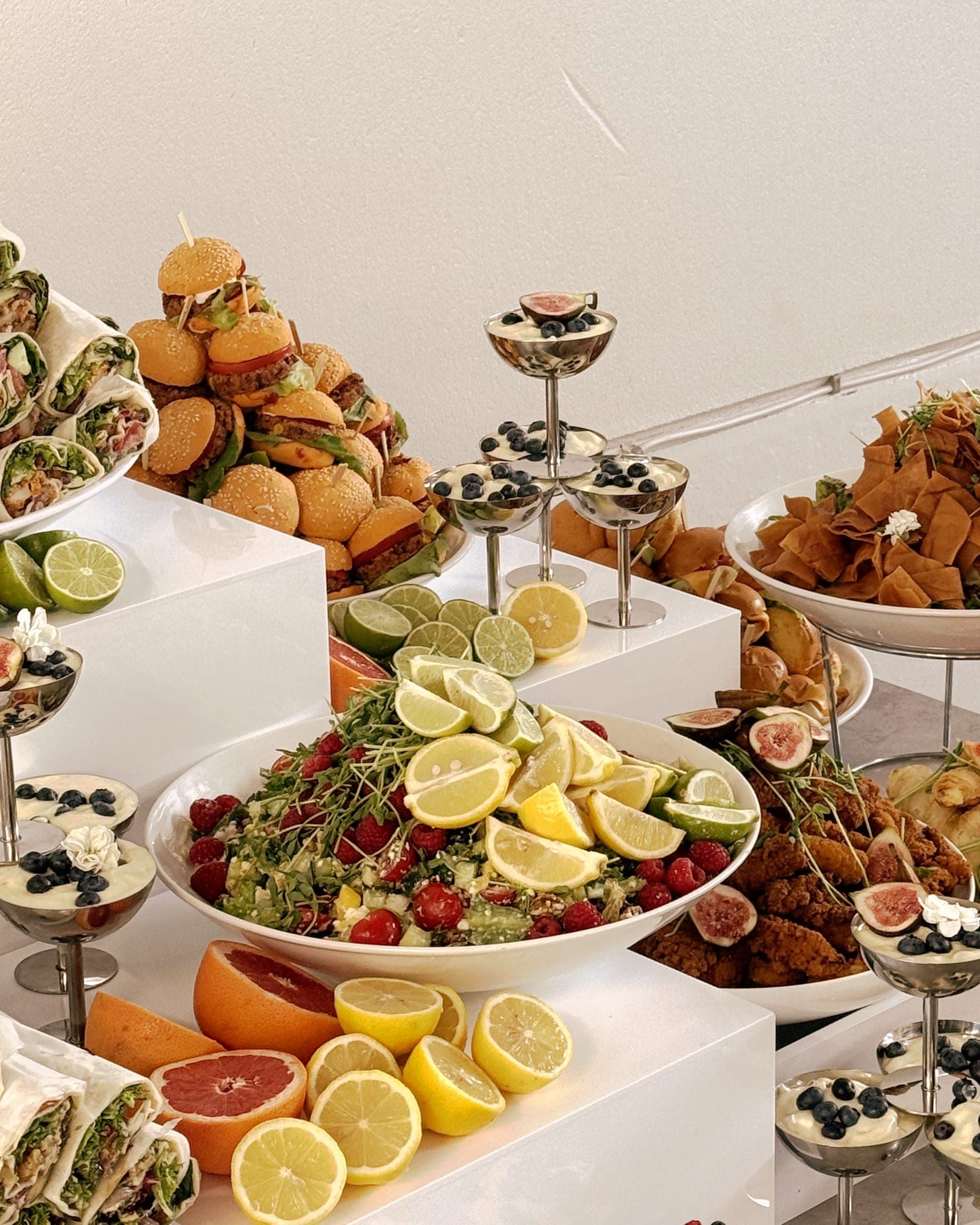 Nahaufnahme: Kräutersalat mit Zitronenscheiben und Feigen in einer weißen Schale, dahinter Mini-Burger, Wraps und Dessertgläser