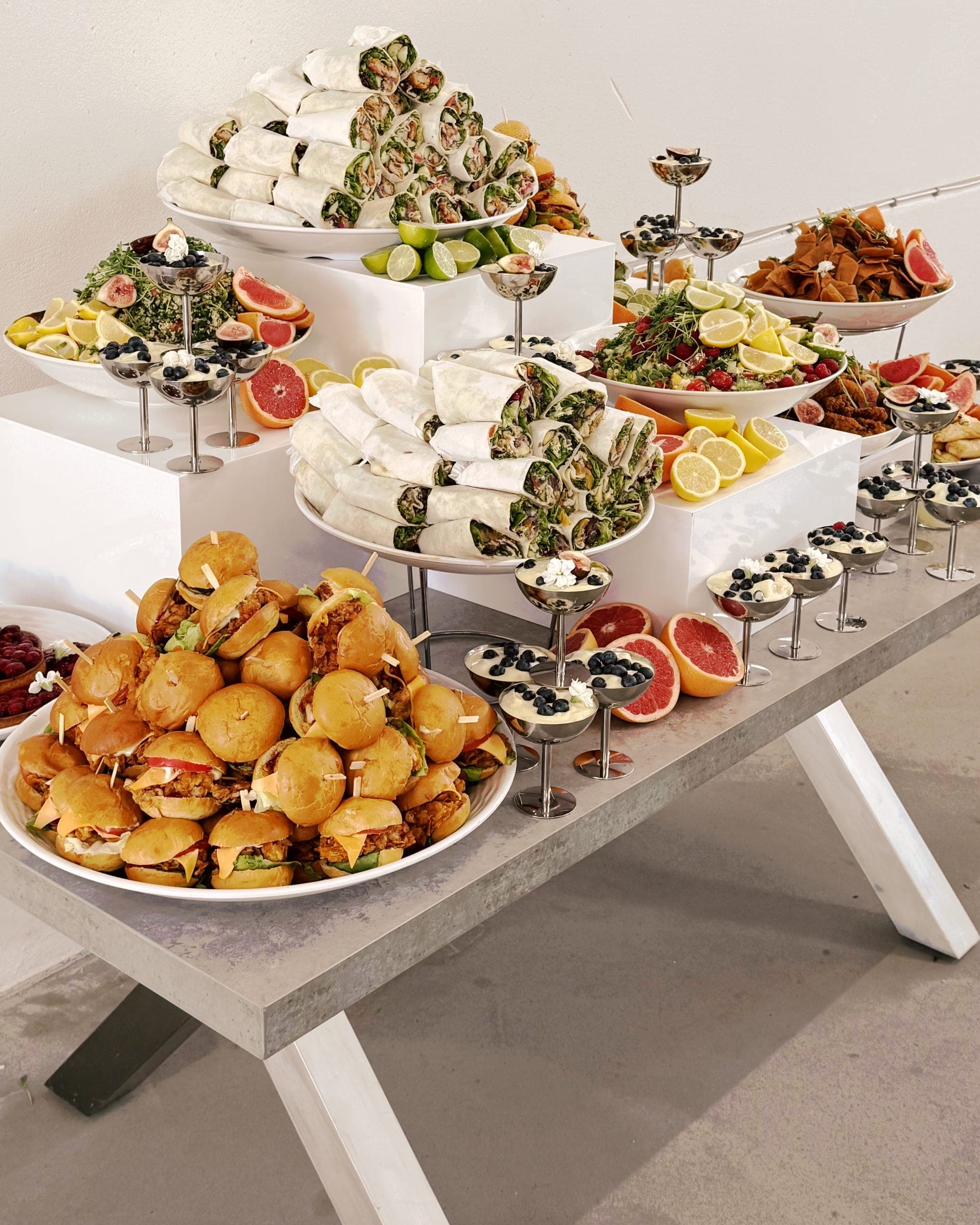 Buffettisch für ein Firmenevent von der Seite: Wraps und Mini-Burger auf Etageren, Salatschalen, Limetten und Dessertgläser auf schmalen Ständern