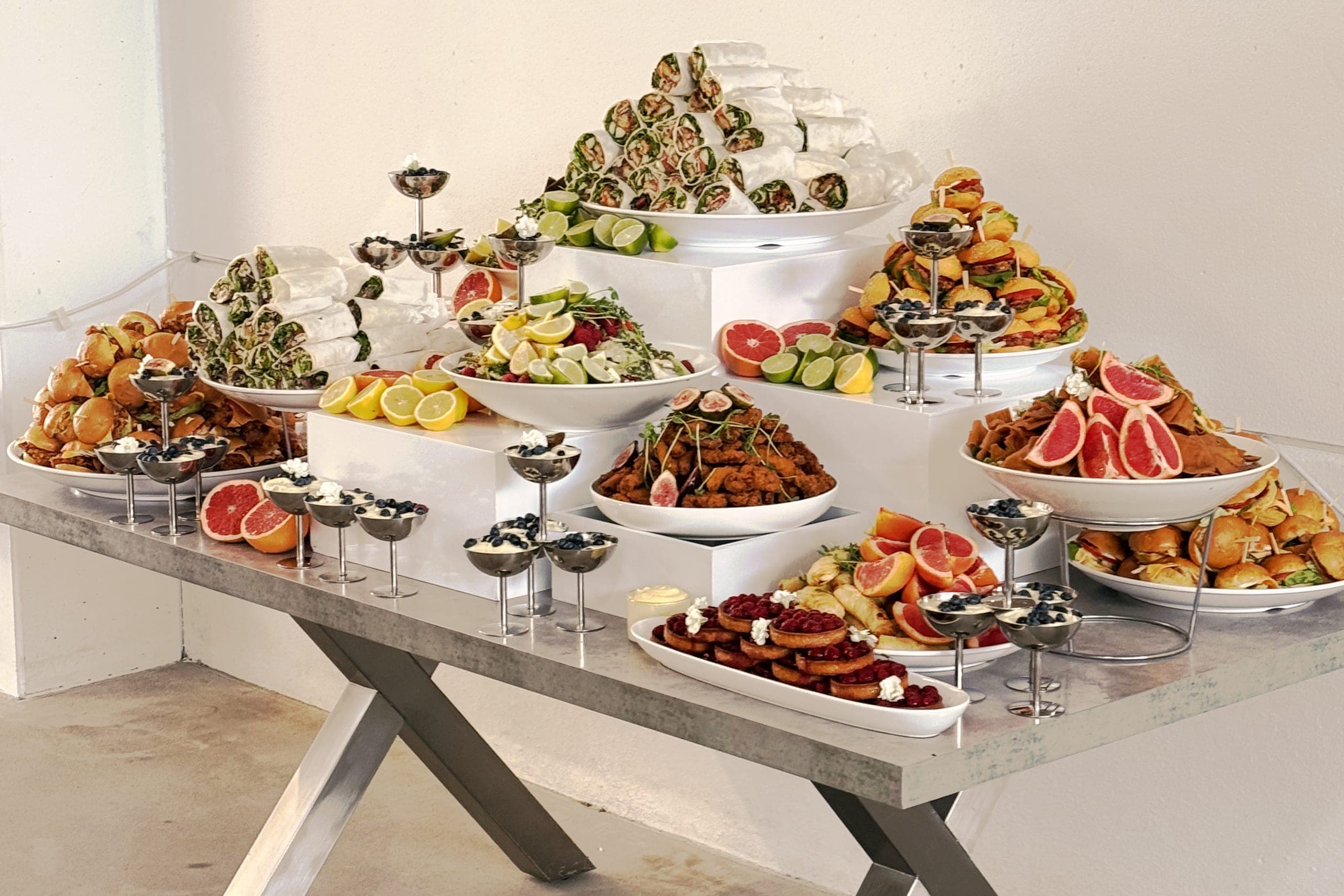 Buffettisch für ein Firmenevent von links: Wraps, Mini-Burger, Salate und Dessertgläser auf weißen Sockeln, davor die gekreuzten Tischbeine
