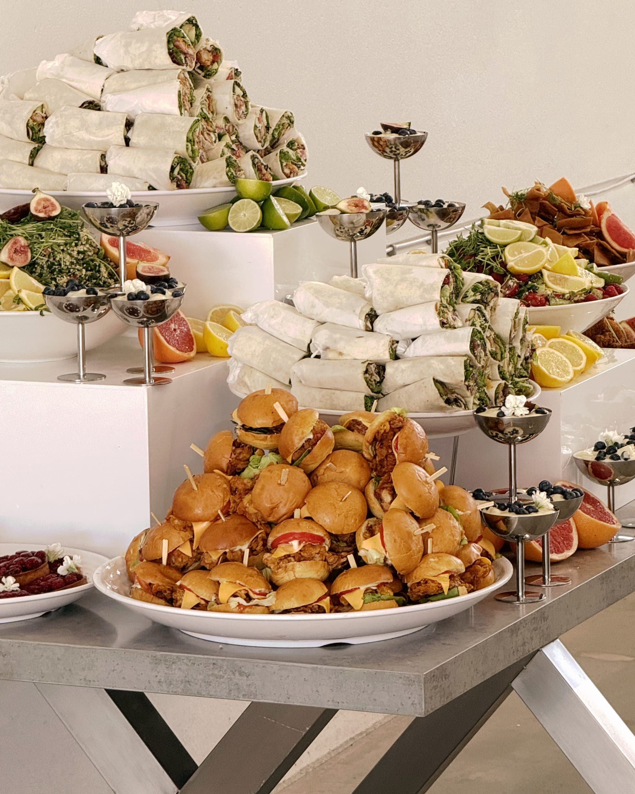 Nahaufnahme eines Buffets: aufgetürmte Wraps mit Salat und Hähnchen, Mini-Burger, Limettenhälften, Dessertgläser mit Blaubeeren und Feigen