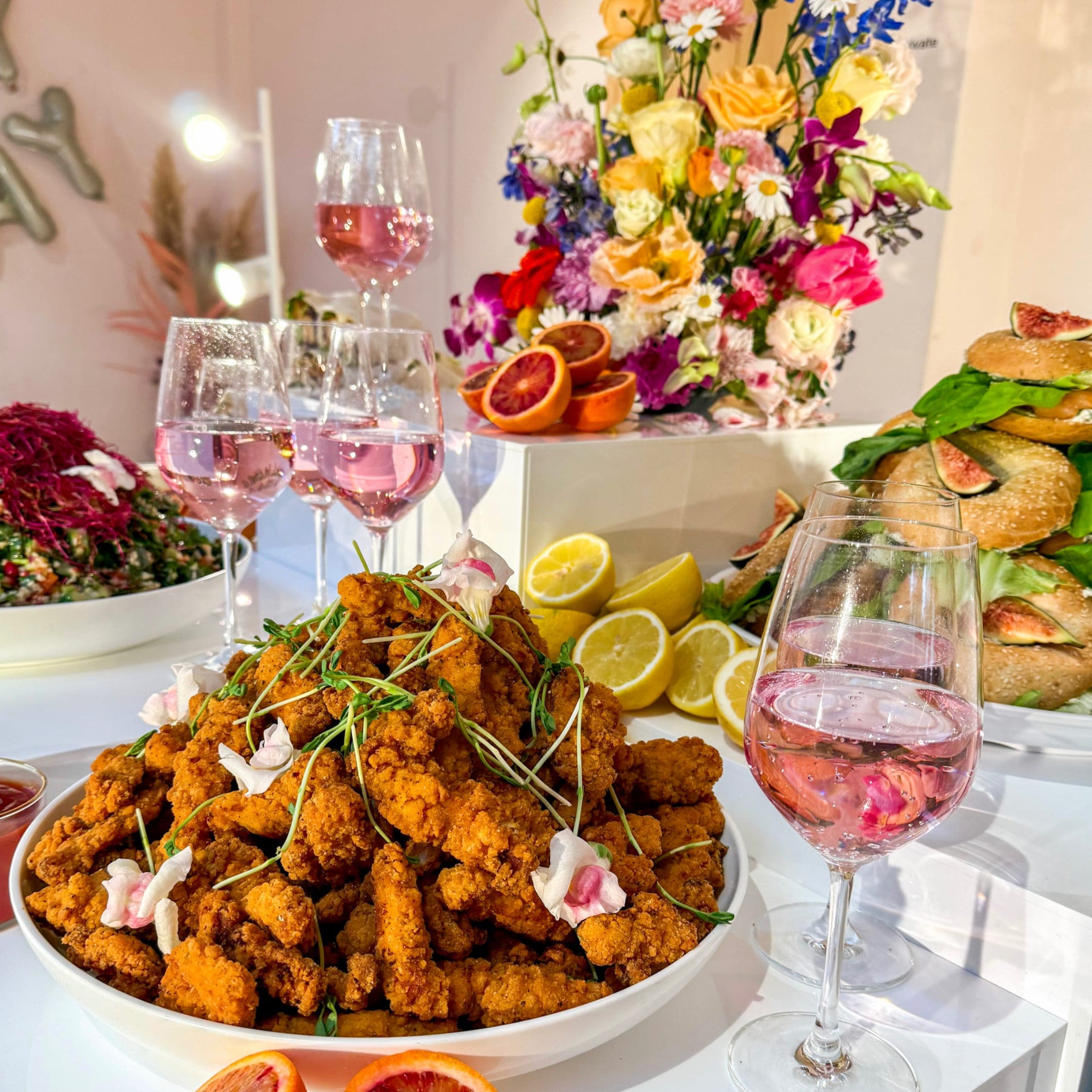 Schale mit knusprigen Hähnchenstreifen und Blüten, dahinter Bagel-Sandwiches mit Feigen und Gläser Rosé