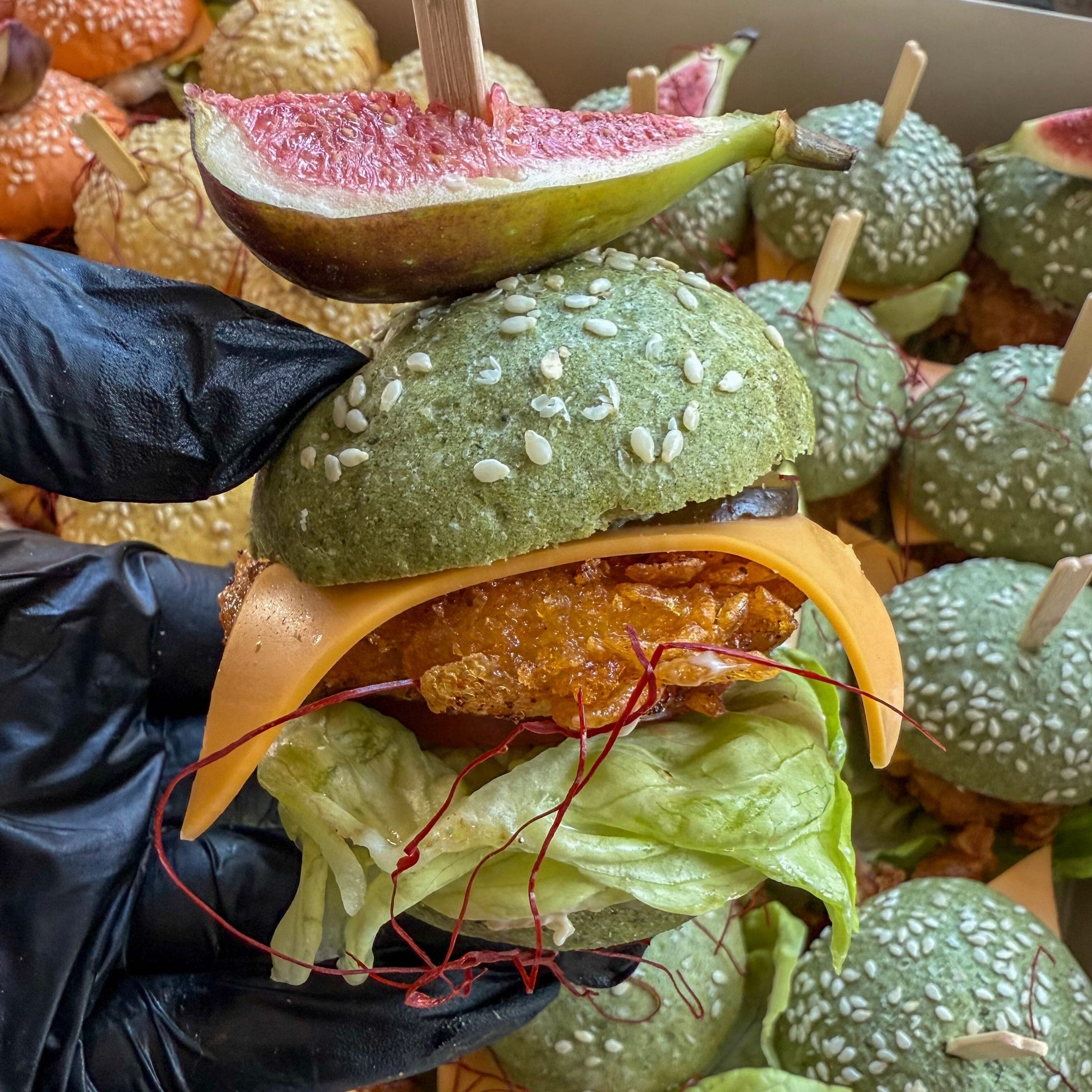 Mini-Burger im grünen Sesambrötchen mit Käse, knusprigem Hähnchen, Salat und einer halben Feige auf dem Spieß