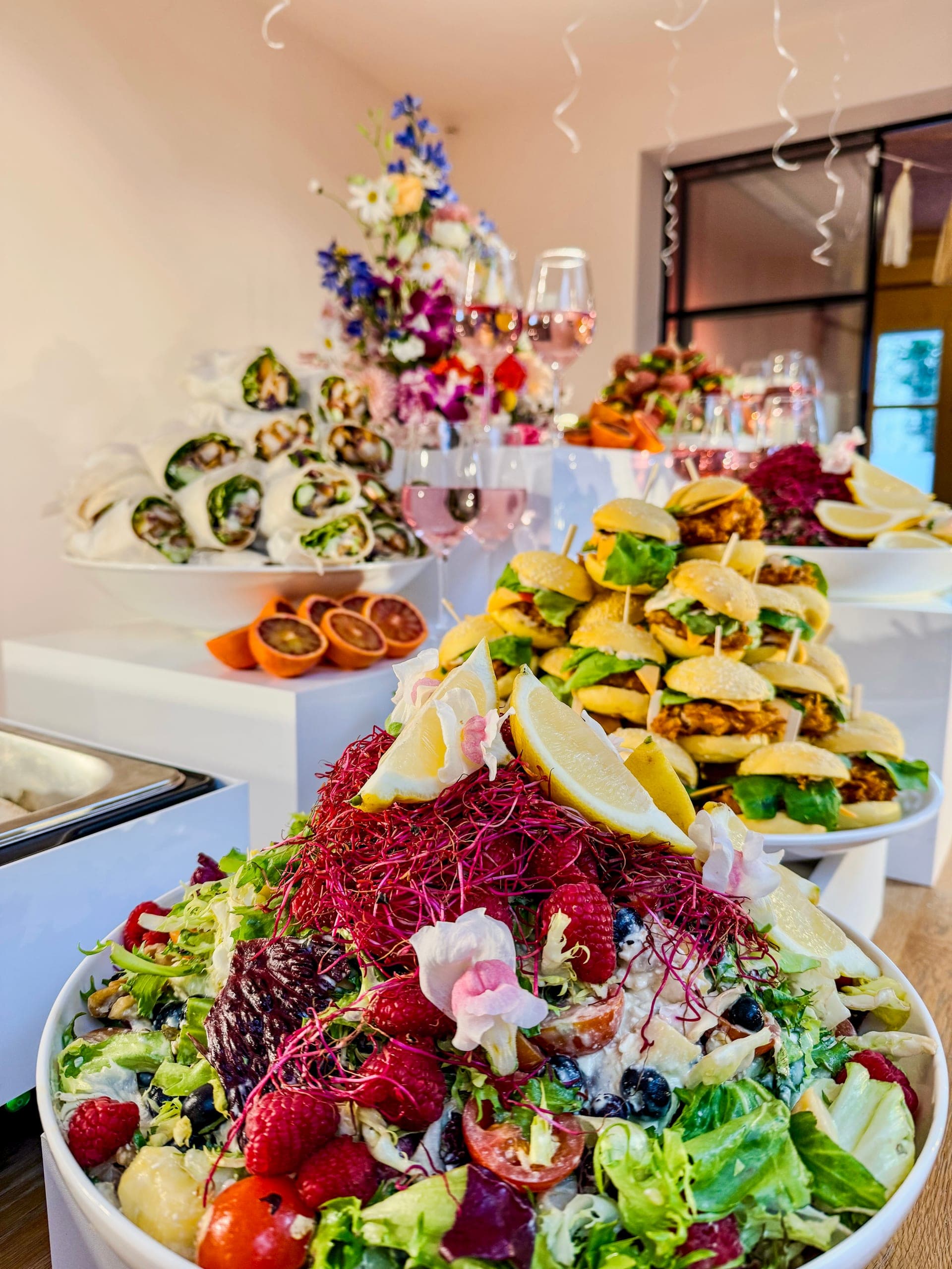 Buffet mit großer Salatschale mit Beeren und Blüten, Mini-Burgern auf einem Podest gestapelt und Wrap-Röllchen auf Risern