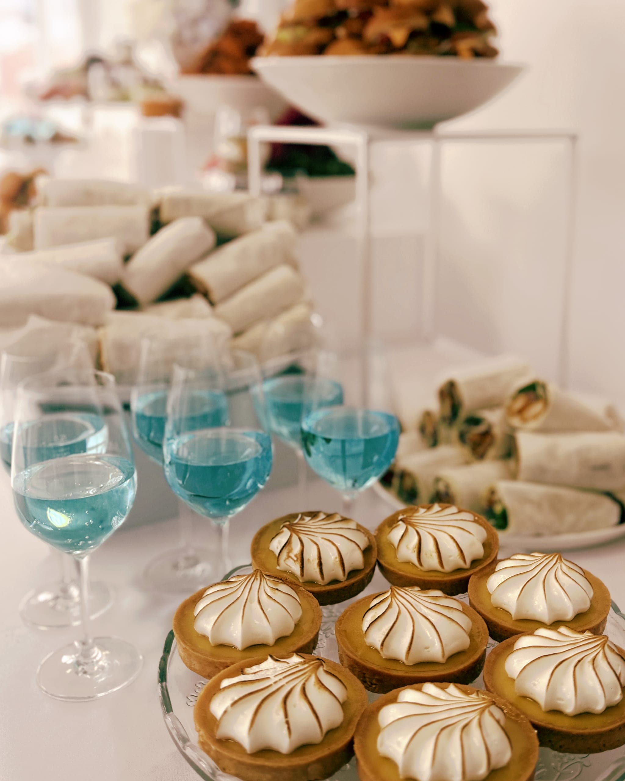 Blaue Cocktails in Weingläsern neben Baiser-Tartelettes und einer Platte gerollter Wraps auf weißer Tischdecke