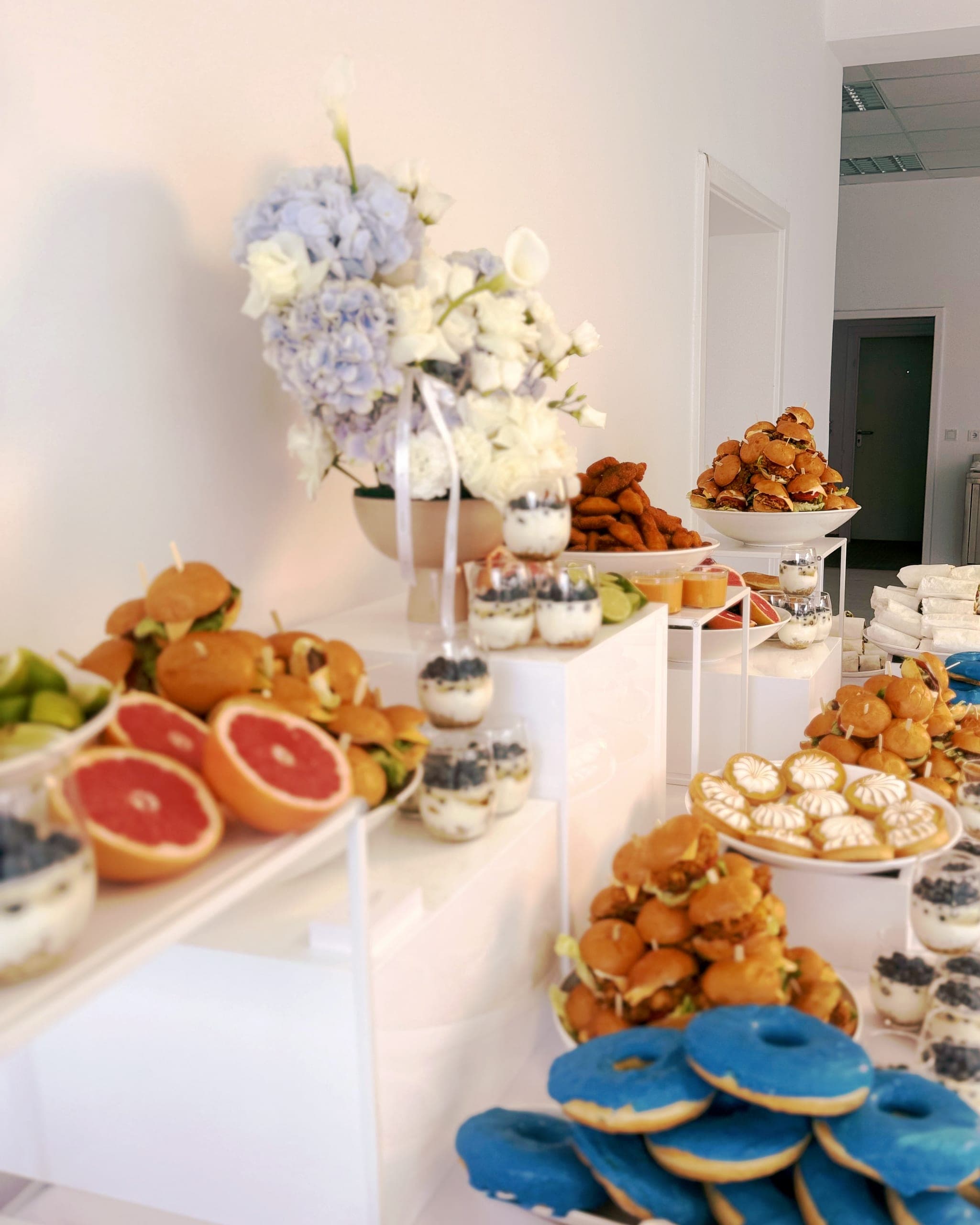 Strauß aus blauen Hortensien und weißen Lilien über einem Buffet mit Grapefruithälften, Mini-Burgern und Dessertgläsern mit Blaubeeren