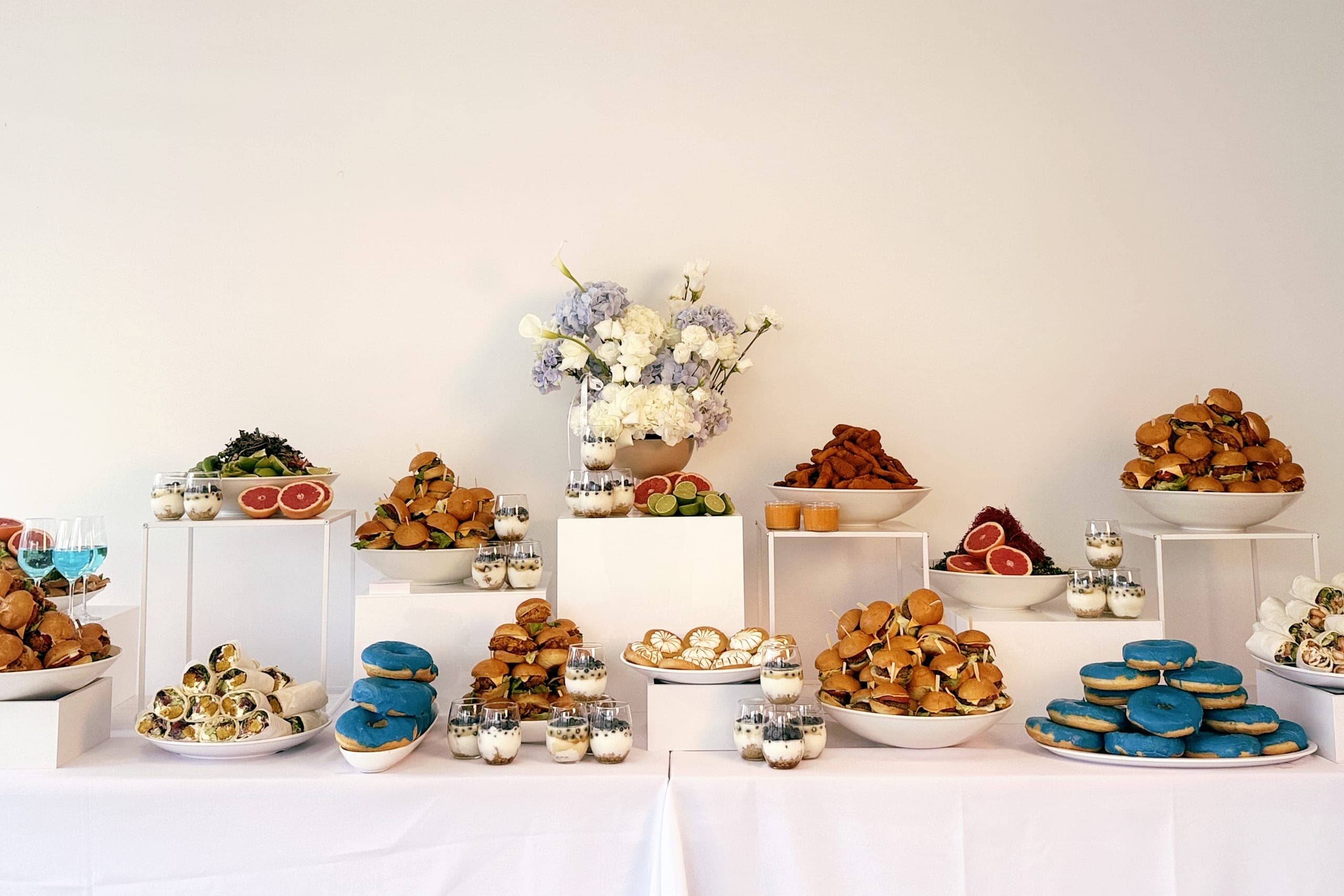 Buffet für ein Firmenevent auf weißen Sockeln: Mini-Burger, Wraps, blaue Donuts, Tartelettes und Dessertgläser, ein Blumenstrauß in Blau und Weiß