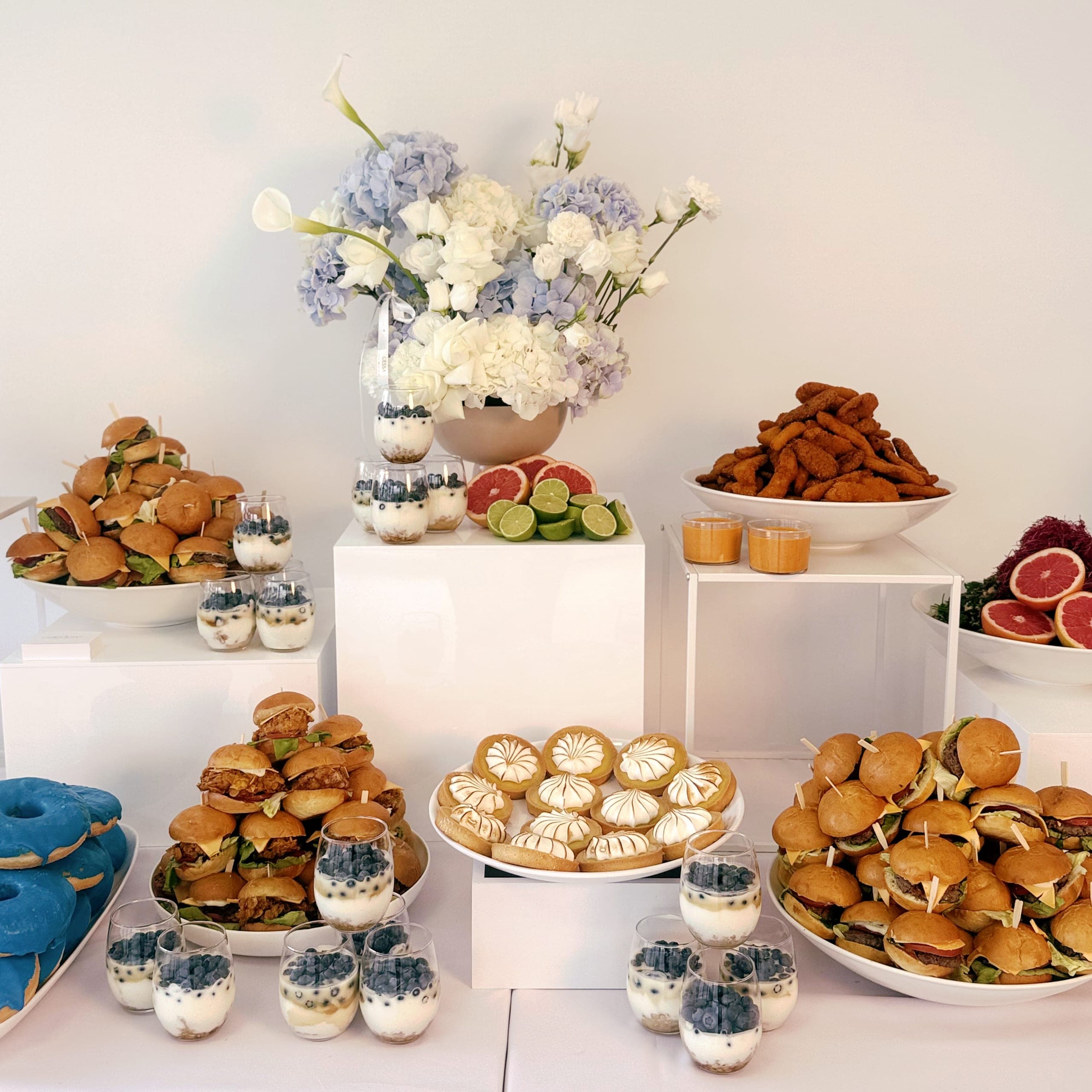 Buffet für ein Firmenevent frontal: Blumen in Blau und Weiß über Mini-Burgern, Baiser-Tartelettes, Dessertgläsern mit Blaubeeren, Grapefruit und einem blauen Donut
