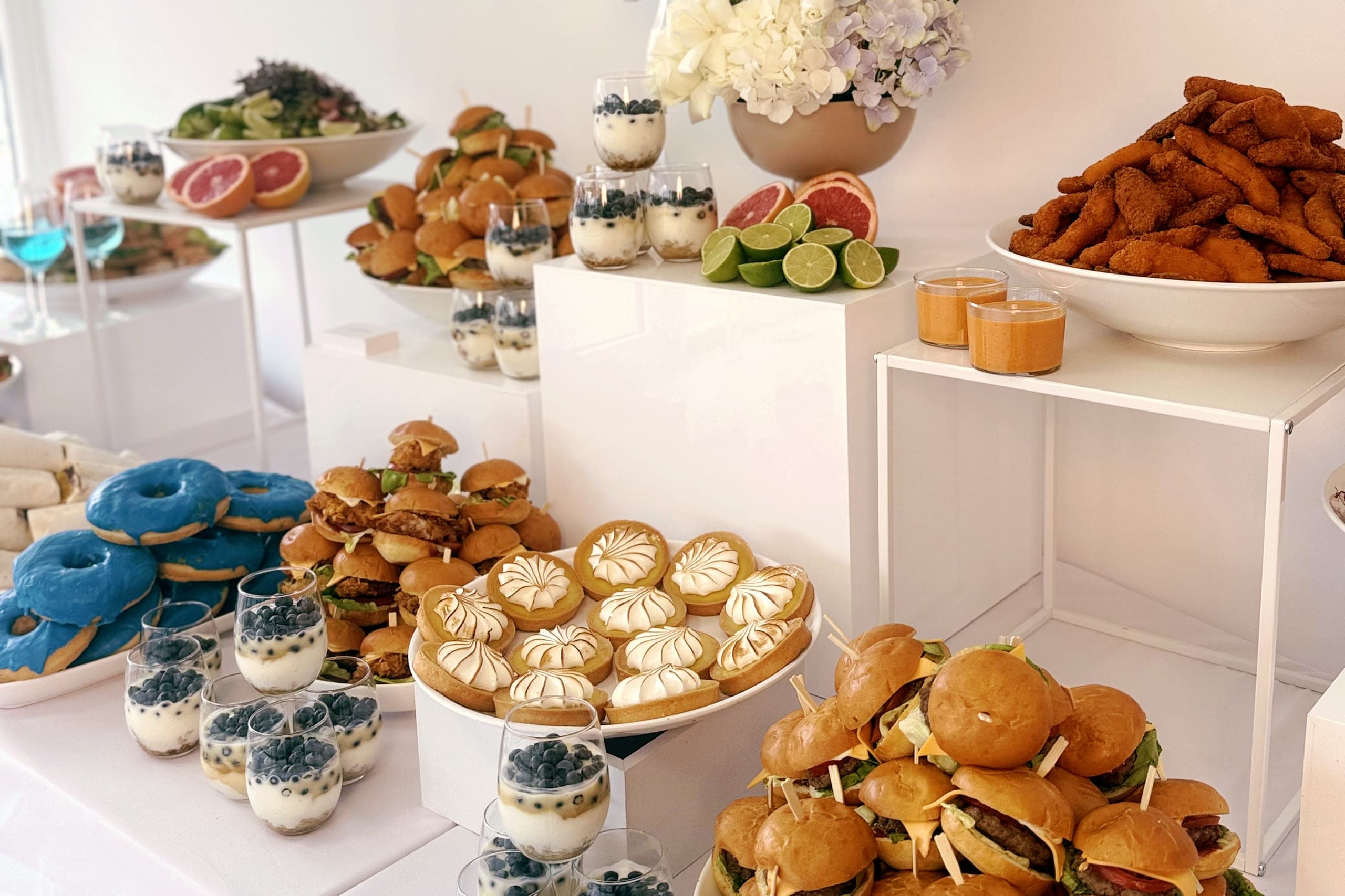 Langes Buffet in einem hellen Raum: Blumen in Blau und Weiß, Grapefruit, Limetten, Mini-Burger, blaue Donuts und Dessertgläser