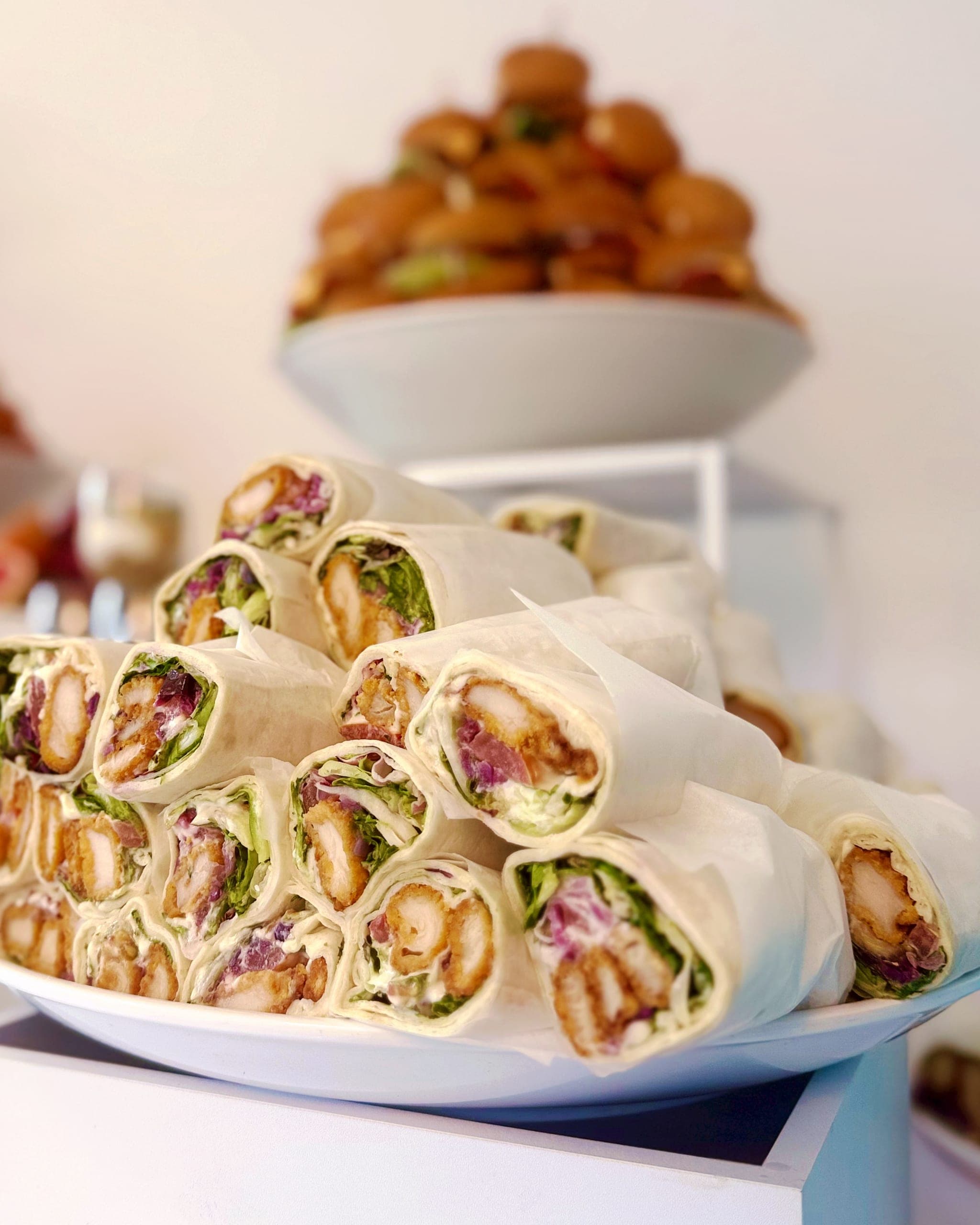 Nahaufnahme gerollter Wraps mit Hähnchen, Salat und Rotkohl auf einem weißen Sockel, dahinter Mini-Burger und blaue Donuts