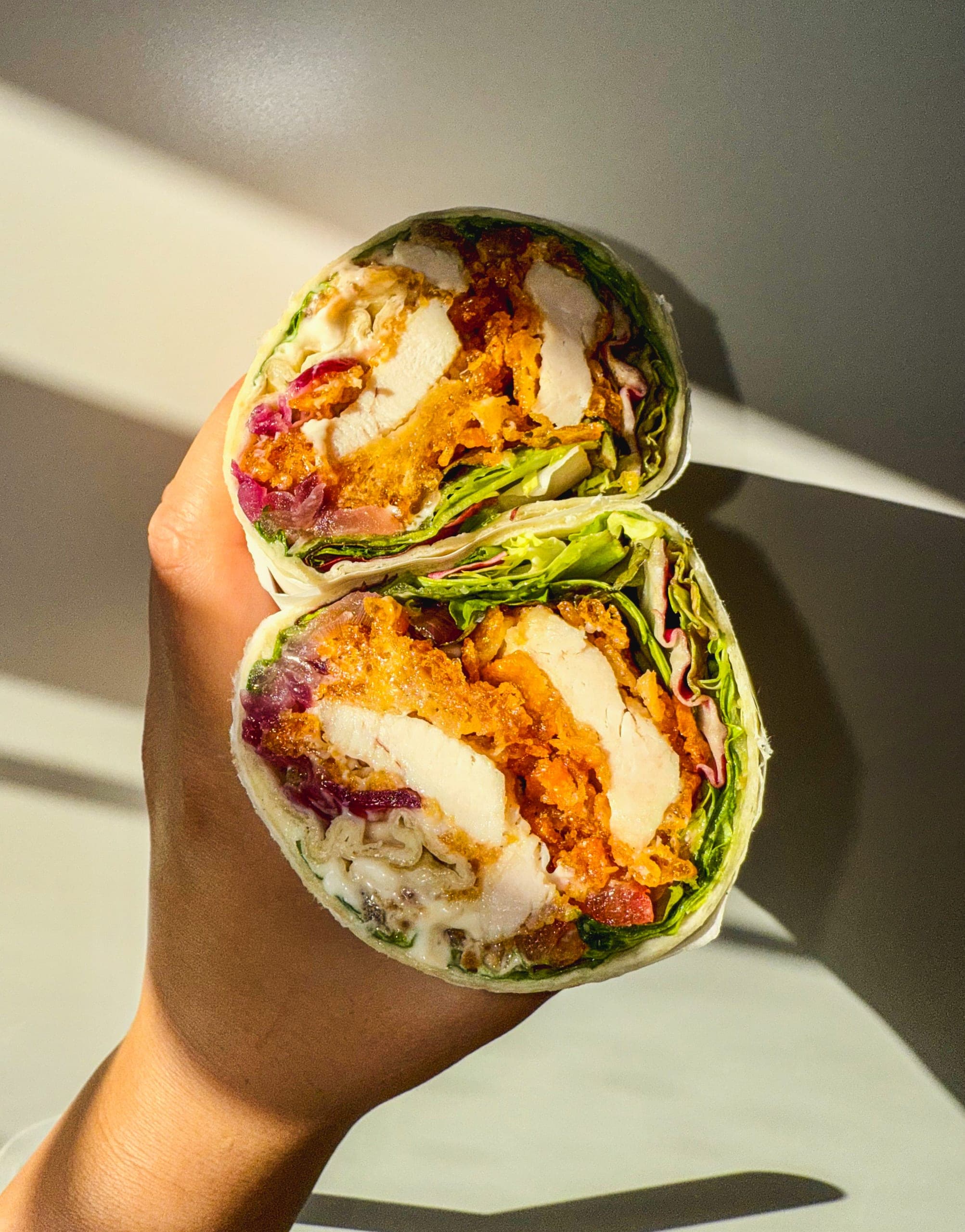 Aufgeschnittener Wrap mit Hähnchenbrust, Panade, Rotkohl und Salat, im harten Sonnenlicht in der Hand gehalten